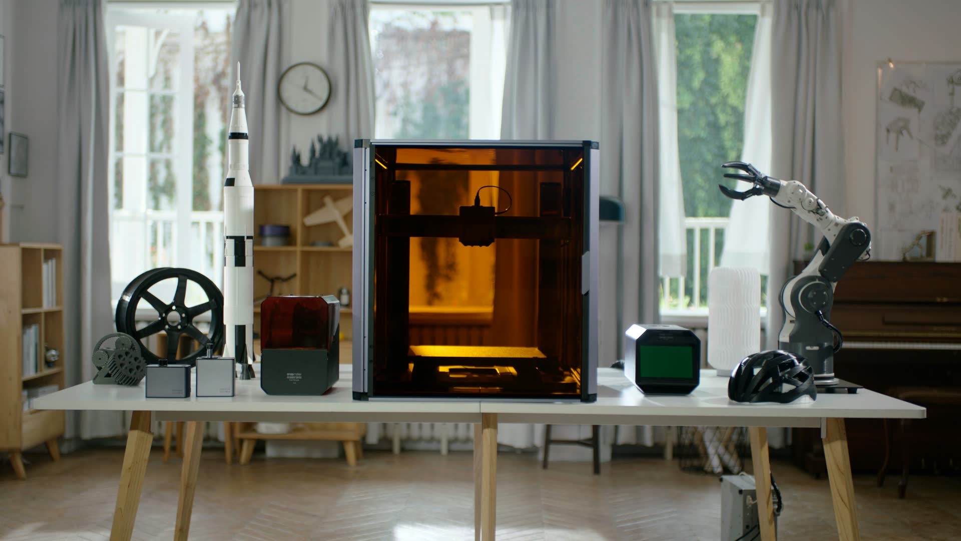 A 3D printer on a workbench, surrounded by printed objects: a wheel, a rocket, a helmet and a robot arm